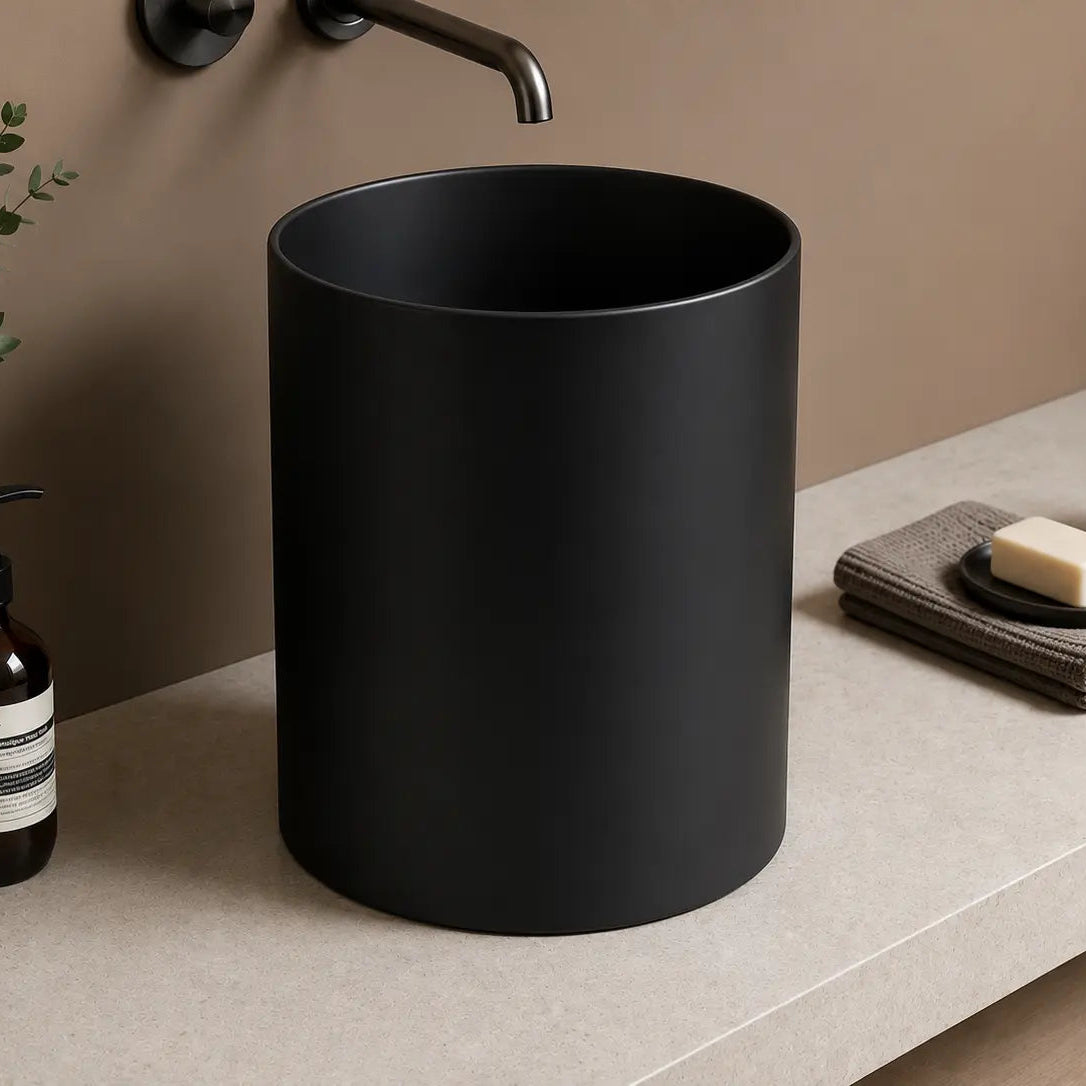 Black cylindrical trash can on a bathroom counter with soap, towel, and candle.